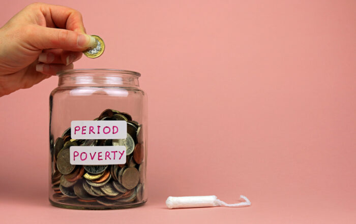 A hand dropping a coin into a glass jar filled with money, labeled with stickers that read "PERIOD POVERTY." A single white tampon lies on the pink background next to the jar.