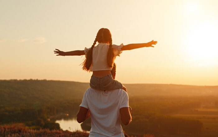 A young girl sitting on her father's shoulders with her arms wide open against a sunset sky.